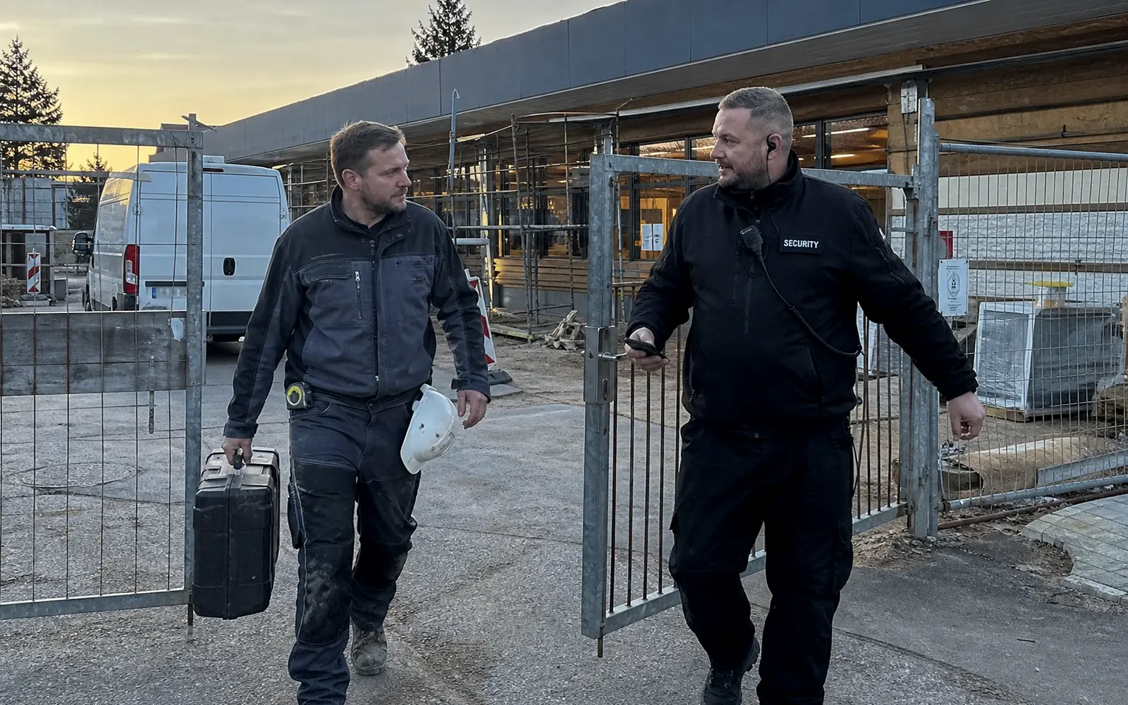 Handwerker mit Werkzeugkoffer und Sicherheitsmitarbeiter am Tor einer Baustelle